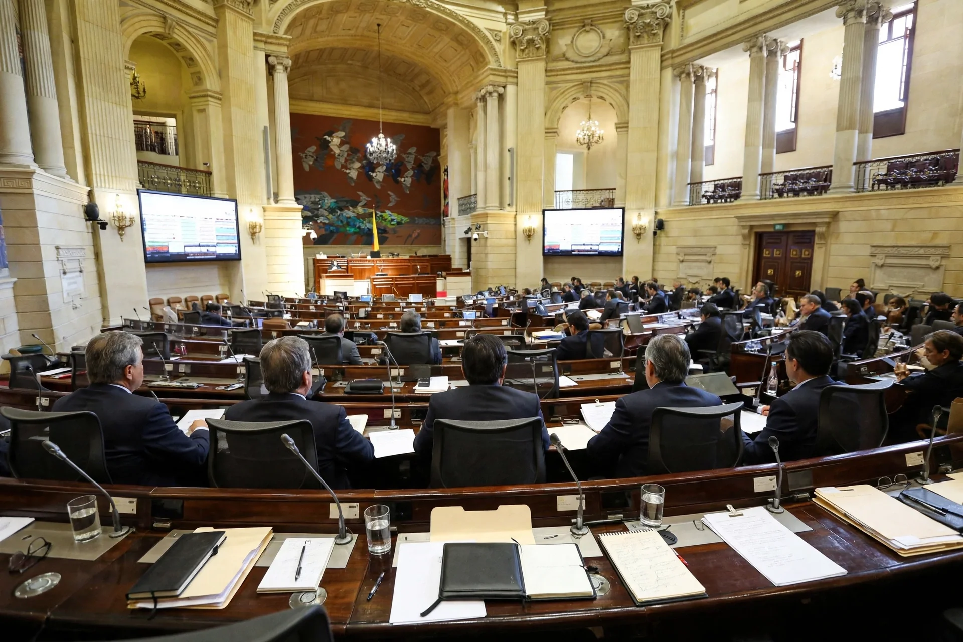 Recinto del Senado de Colombia visto desde los escaños, con senadores de espaldas en una mesa curva de madera con documentos y micrófonos