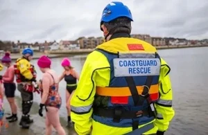 A Coastguard Rescue Officer in a yellow jacket standing on a beach near swimmers
