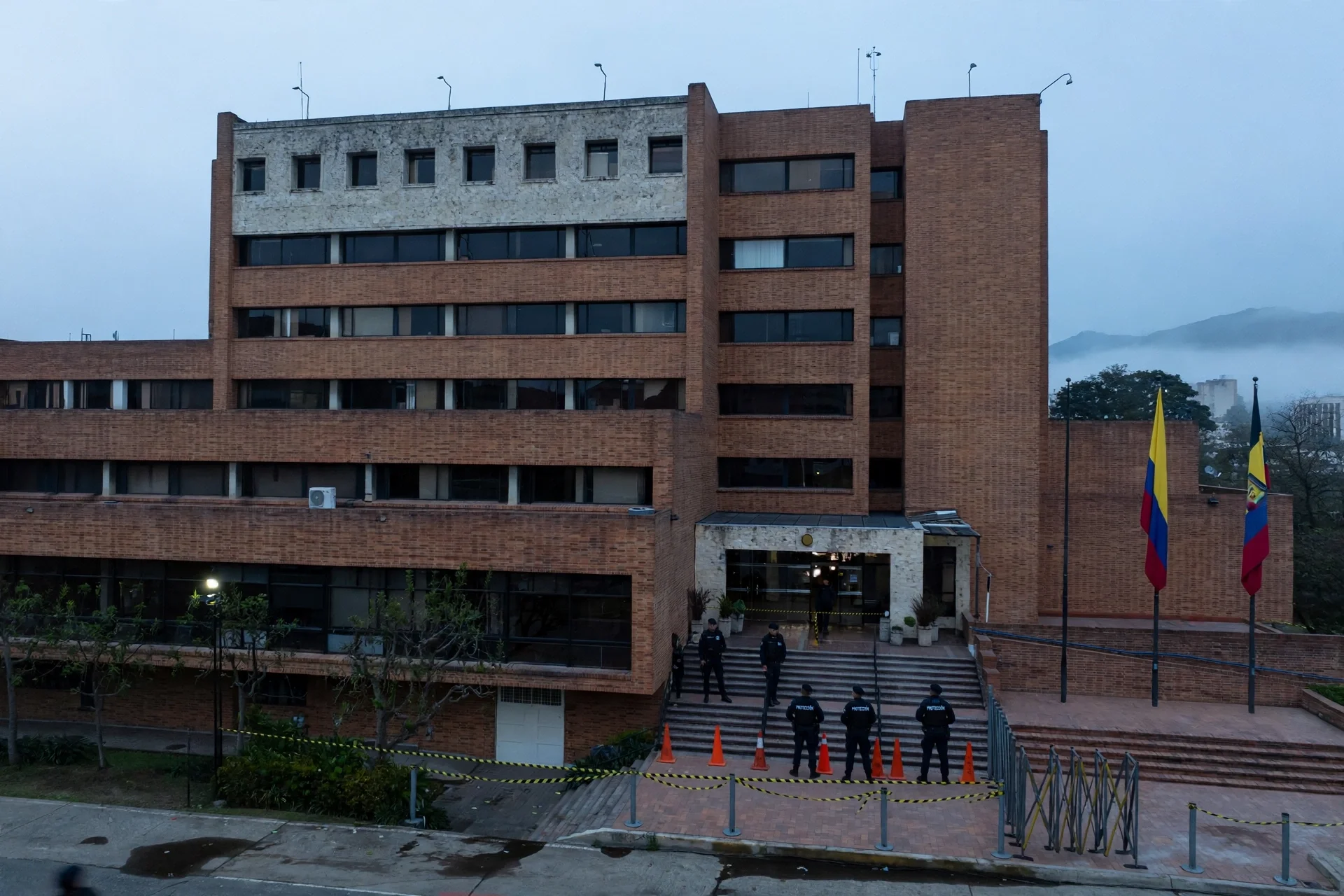Edificio institucional de ladrillo al amanecer con cordón de seguridad, conos y agentes de protección en la escalinata de entrada y banderas de Colombia