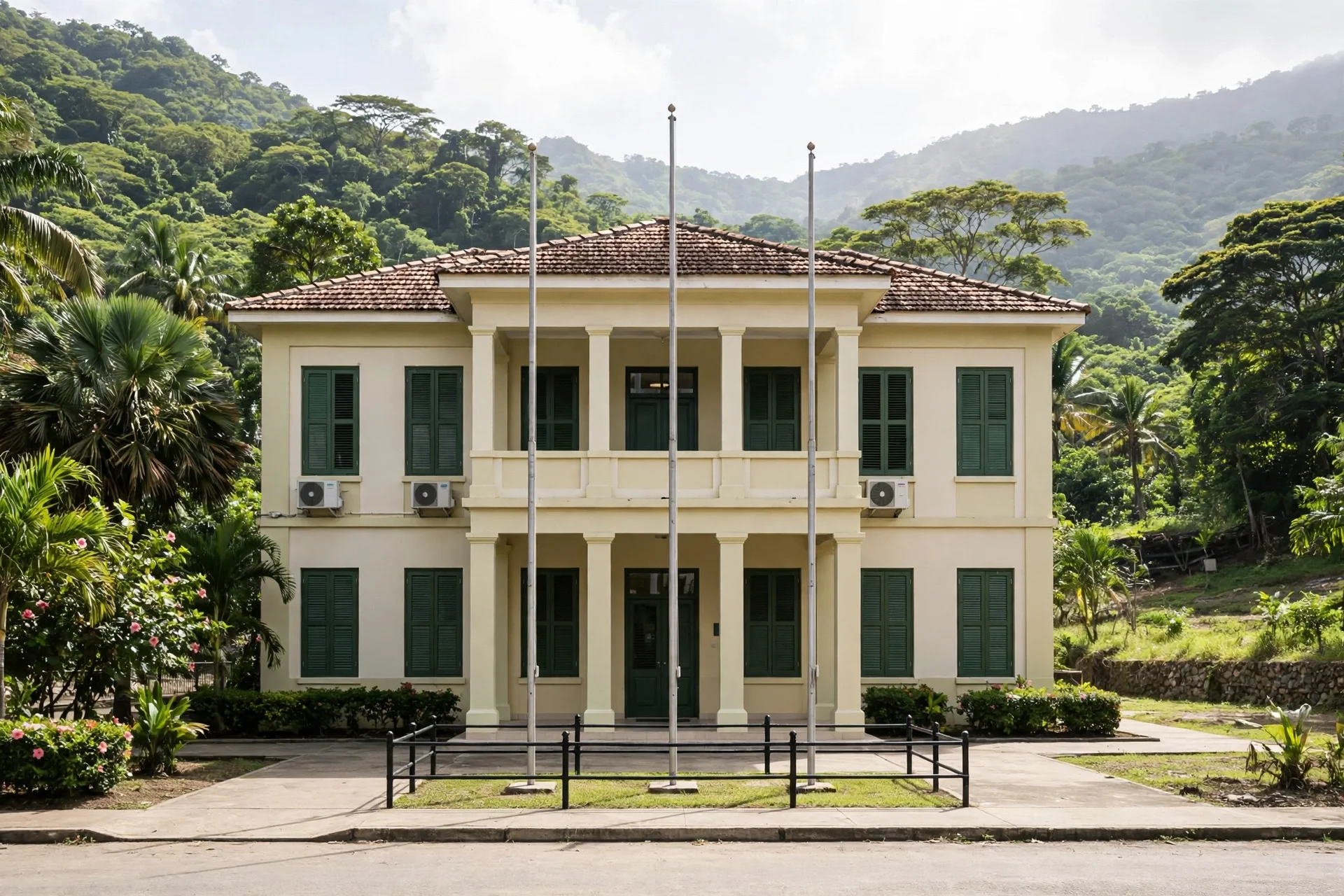 Edificio gubernamental caribeño de dos plantas con mástiles sin banderas frente a montañas verdes