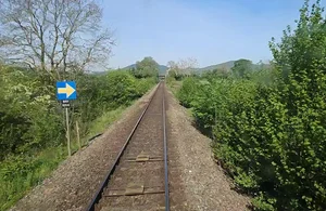 Single railway track stretching ahead through green countryside, with a blue sign bearing a yellow arrow on the left