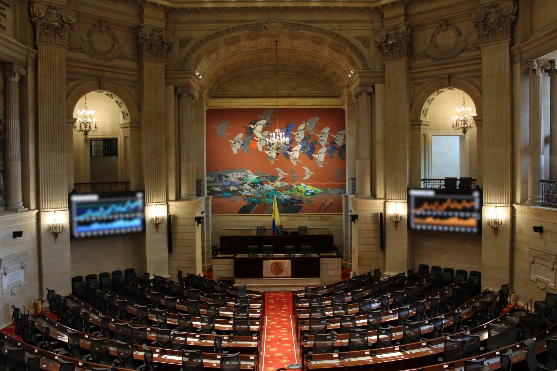 Hemiciclo vacío del Congreso colombiano con pantallas laterales que muestran gráficos financieros desenfocados en un ambiente institucional