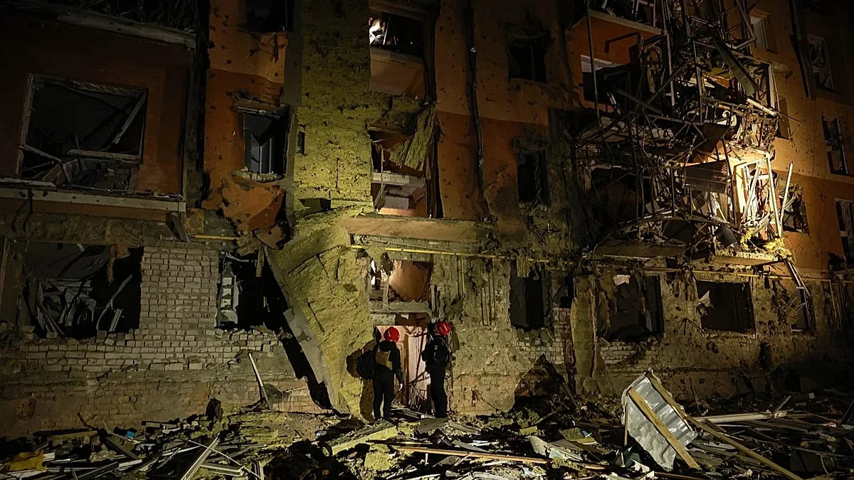 Dos rescatistas con cascos rojos frente a un edificio residencial destruido por un ataque aéreo, entre escombros, durante la noche en Zaporiyia