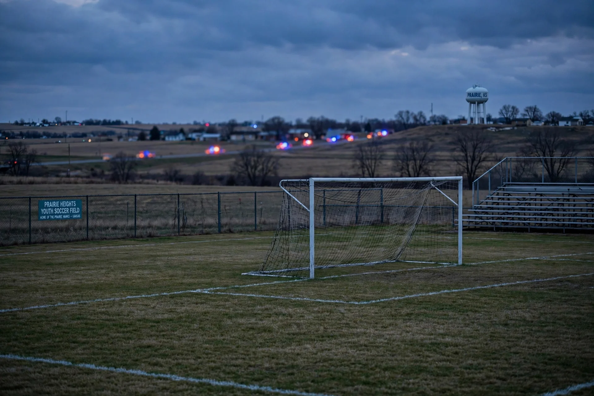 Cancha juvenil de fútbol vacía al anochecer, con la portería en primer plano y luces lejanas.